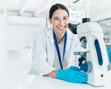 Eine Wissenschaftlerin in einem Labor lächelt, während sie an einem Mikroskop arbeitet. Sie trägt einen weißen Laborkittel und Handschuhe.