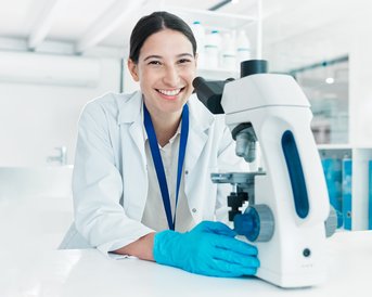 Eine Wissenschaftlerin in einem Labor lächelt, während sie an einem Mikroskop arbeitet. Sie trägt einen weißen Laborkittel und Handschuhe.