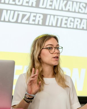 Eine Frau mit Brille spricht während einer Präsentation. Im Hintergrund sind Texte zu sehen, die "Neuer Denkansatz" und "Nutzer Integration" beinhalten. Ein Laptop mit dem Apple-Logo steht auf dem Tisch vor ihr.