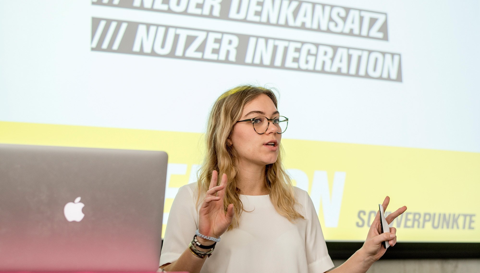 Eine Frau mit Brille spricht während einer Präsentation. Im Hintergrund sind Texte zu sehen, die "Neuer Denkansatz" und "Nutzer Integration" beinhalten. Ein Laptop mit dem Apple-Logo steht auf dem Tisch vor ihr.