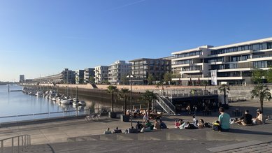 Moderne Wohngebäude am Wasser mit Booten im Hafen. Menschen sitzen auf Stufen und genießen die Sonne. Klarer blauer Himmel.