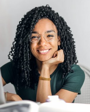 Eine lächelnde Frau mit lockigem Haar sitzt an einem Tisch, gestützt auf ihre Hand. Vor ihr steht ein Laptop, während sie in die Kamera blickt. Der Hintergrund ist hell und neutral.