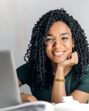 A smiling woman with curly hair sits at a table, resting her hand on it. In front of her is a laptop as she looks into the camera. The background is bright and neutral.