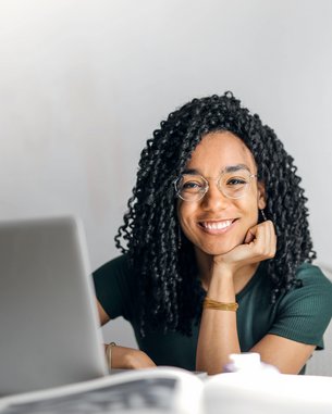 Eine lächelnde Frau mit lockigem Haar sitzt an einem Tisch, gestützt auf ihre Hand. Vor ihr steht ein Laptop, während sie in die Kamera blickt. Der Hintergrund ist hell und neutral.
