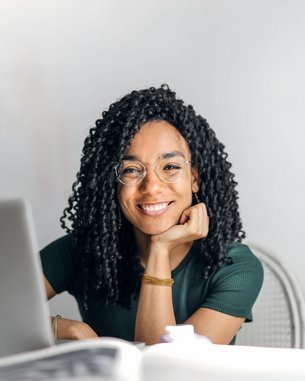 A smiling woman with curly hair sits at a table, resting her hand on it. In front of her is a laptop as she looks into the camera. The background is bright and neutral.