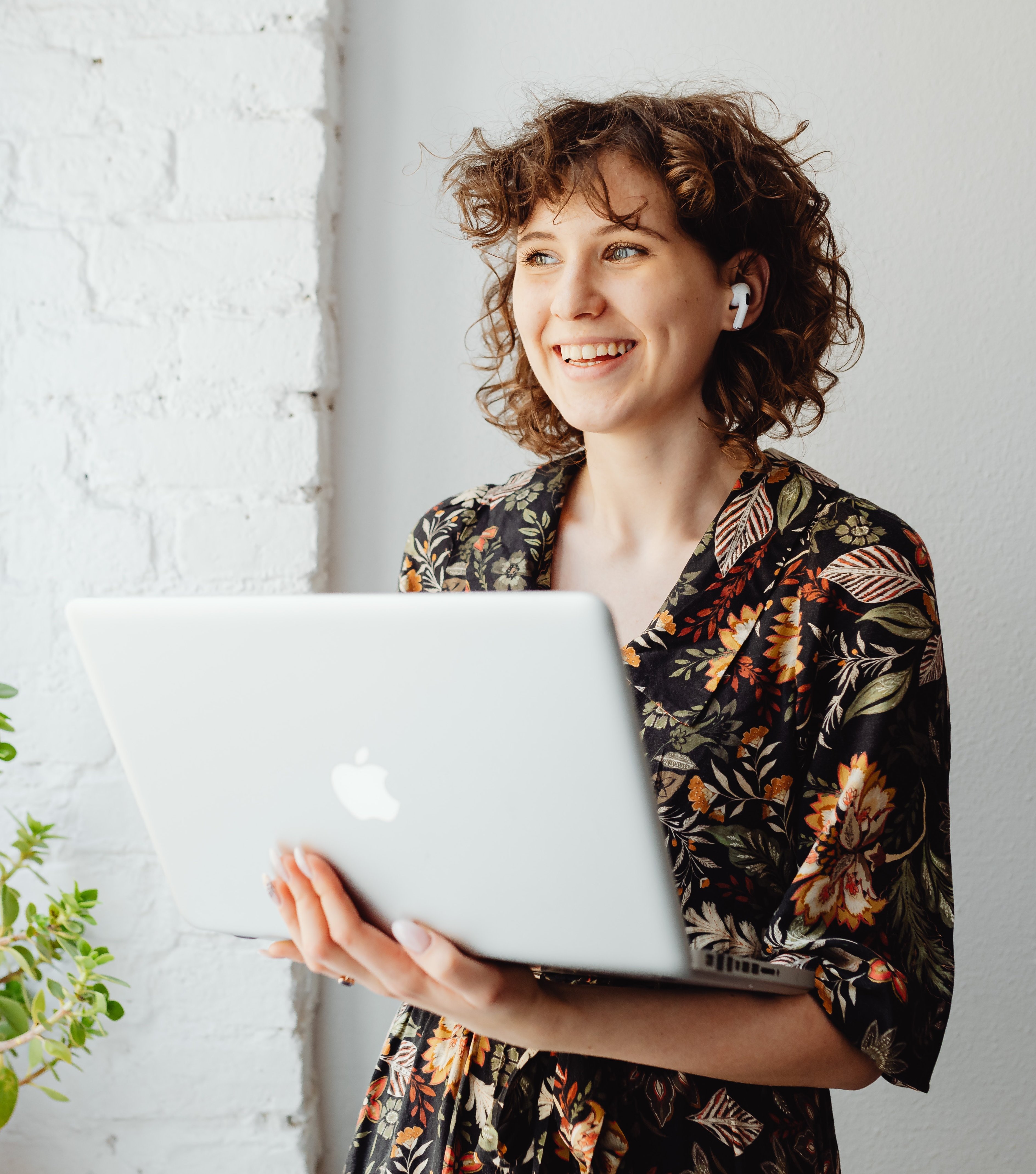 Eine Frau mit lockigem Haar steht neben einer Pflanze und hält einen Laptop. Sie trägt ein blumiges Oberteil und hat kabellose Ohrhörer im Ohr. Der Hintergrund ist eine helle, gemauerte Wand.