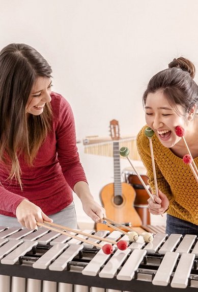 Two women are playing a xylophone together and laughing. In the background, various musical instruments can be seen.