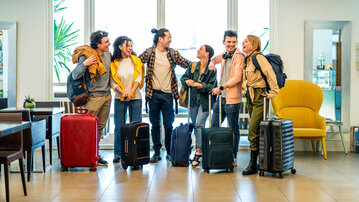 A group of seven people stands in a bright room with luggage. They are smiling and chatting as they look towards the exit.