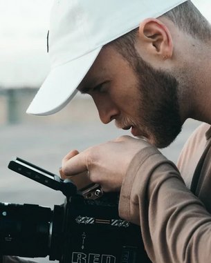 Ein Mann mit einer weißen Kappe betrachtet sorgfältig eine Kamera. Er hält die Kamera mit einer Hand und justiert sie mit der anderen. Der Fokus liegt auf seiner Konzentration und der professionellen Ausrüstung.