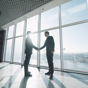 Two men in suits shake hands in front of large windows, through which sunlight streams.