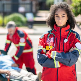 Eine Rettungssanitäterin in rot-gelber Uniform steht mit verschränkten Armen im Vordergrund, während Kollegen einen Patienten auf einer Trage behandeln.