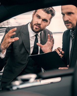 Zwei Männer in Anzügen diskutieren im Innenraum eines Autos. Einer zeigt mit den Händen auf etwas, während der andere Notizen auf einem Tablet macht. Beide scheinen an einem geschäftlichen Gespräch beteiligt zu sein.