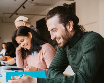 A group of people is sitting at a table, working on documents. A man with long hair and a green sweater is writing, while a woman in a pink sweater smiles and takes notes as well.