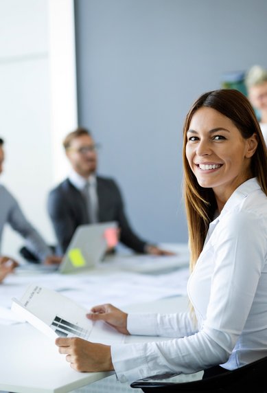 Eine Frau in einem weißen Hemd lächelt und hält Dokumente, während sie an einem Konferenztisch mit mehreren Personen in einem Besprechungsraum sitzt.