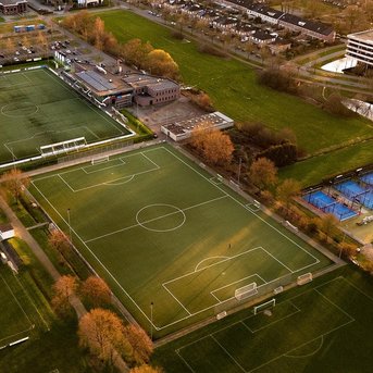 Luftaufnahme eines Sportgeländes mit mehreren Fußballfeldern und Tennisplätzen, umgeben von Bäumen und einem Parkplatz.