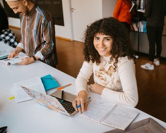 Eine lächelnde Frau mit lockigem Haar sitzt an einem Tisch und arbeitet an einem Laptop. Vor ihr liegen Notizblätter und Stifte. Im Hintergrund sind weitere Personen zu sehen, die ebenfalls an ihren Aufgaben arbeiten.