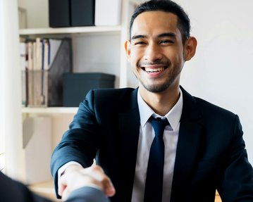 Ein Mann in einem Anzug schüttelt einer anderen Person die Hand und lächelt dabei, während er an einem Tisch sitzt.