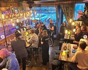Eine belebte Bar mit Gästen an Tischen und der Theke, beleuchtet von warmen Lichtern. Barkeeper serviert Getränke.