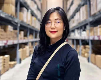 A woman with glasses stands in a warehouse between shelves full of boxes. She is wearing a black top and has long, dark hair.