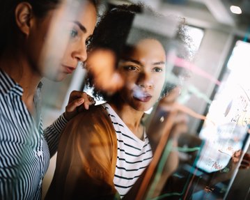 Two women are intently looking at a transparent board filled with diagrams and notes. One woman is pointing at the board while the other watches attentively. Both are in a modern, bright room.