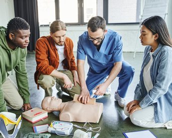 Vier Personen üben Erste Hilfe an einer Übungspuppe, während medizinische Ausrüstung und Materialien um sie herum liegen.