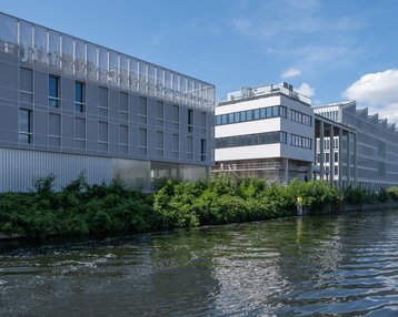 Moderne Gebäude am Wasser mit grüner Vegetation im Vordergrund und einem klaren blauen Himmel im Hintergrund. Die Architektur ist zeitgenössisch und spiegelt eine urbane Umgebung wider.
