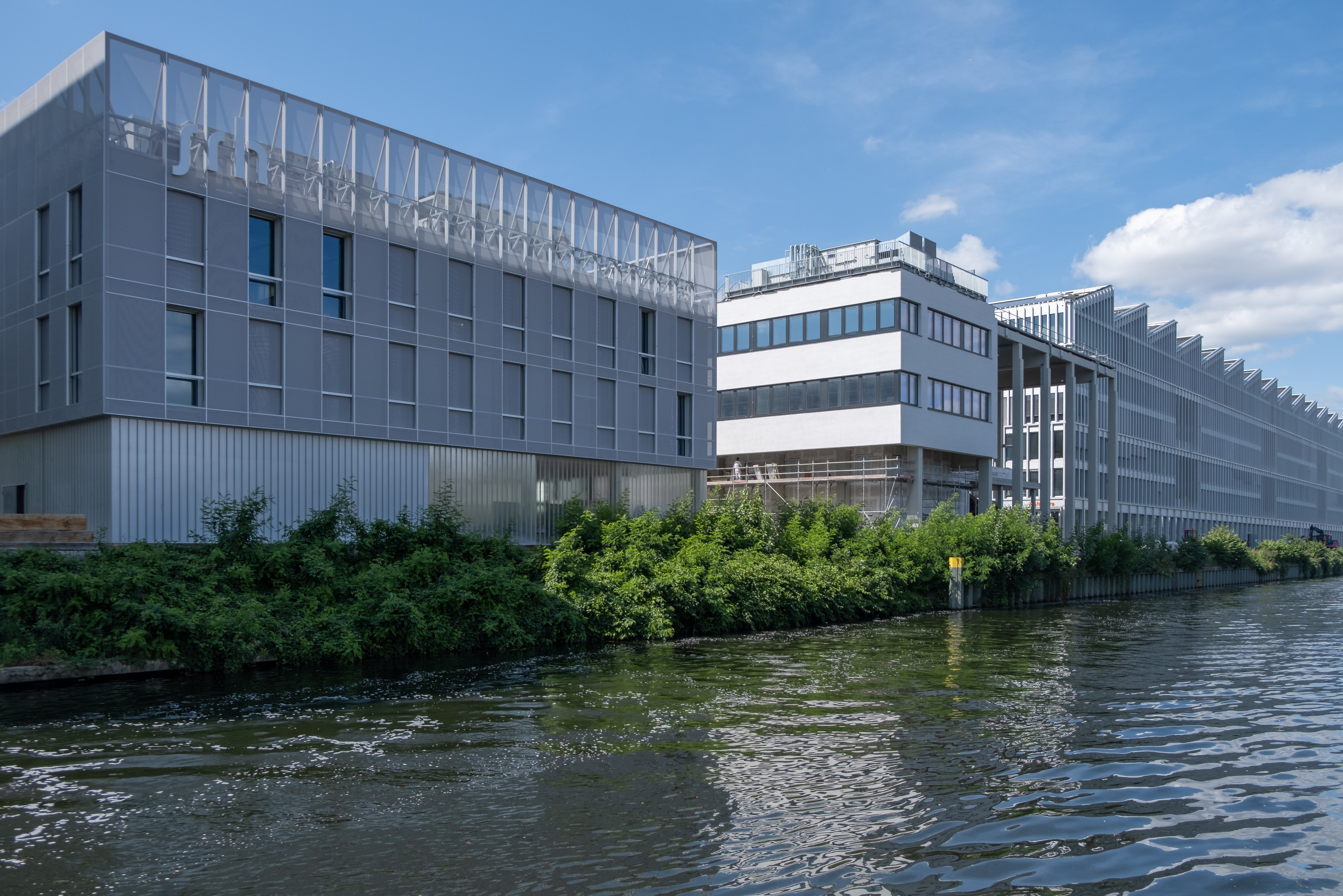 Modern buildings by the water with lush greenery in the foreground and a clear blue sky in the background. The architecture is contemporary and reflects an urban environment.