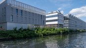 Moderne Gebäude am Wasser mit grüner Vegetation im Vordergrund und einem klaren blauen Himmel im Hintergrund. Die Architektur ist zeitgenössisch und spiegelt eine urbane Umgebung wider.