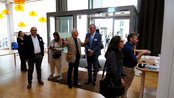 Eine Gruppe von fünf Personen steht in einem modernen Raum. Zwei Männer unterhalten sich, während die anderen auf den Empfangstresen blicken. Helle Lampen hängen von der Decke, und es gibt eine Glas-Tür im Hintergrund.