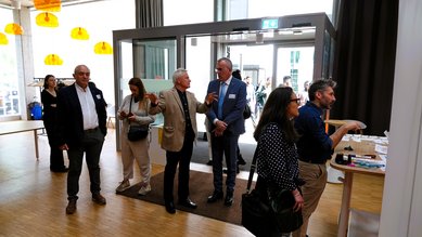 Eine Gruppe von fünf Personen steht in einem modernen Raum. Zwei Männer unterhalten sich, während die anderen auf den Empfangstresen blicken. Helle Lampen hängen von der Decke, und es gibt eine Glas-Tür im Hintergrund.
