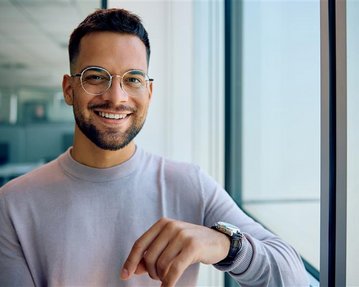 Ein junger Mann mit Brille lächelt und steht neben einem Fenster in einem modernen Büro. Er trägt einen hellen Pullover und eine Armbanduhr.