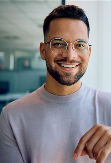 A young man with glasses and a beard smiles as he stands by a window. He is wearing a light sweater and has a watch on his wrist.