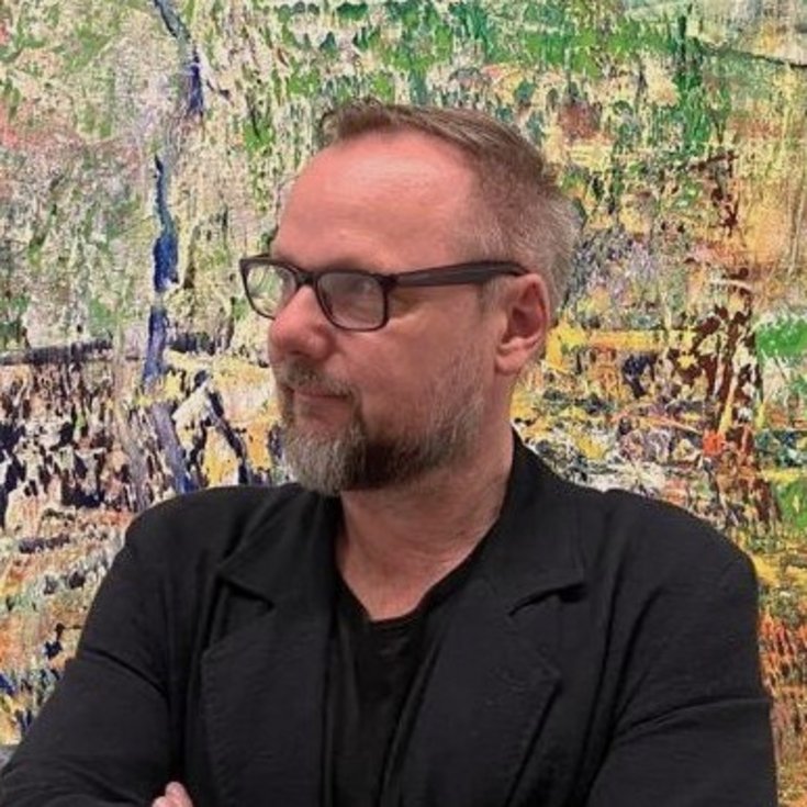Mann mit Brille und Bart, der in einem schwarzen Blazer gekleidet ist, steht vor einer bunten, abstrakten Wandmalerei.