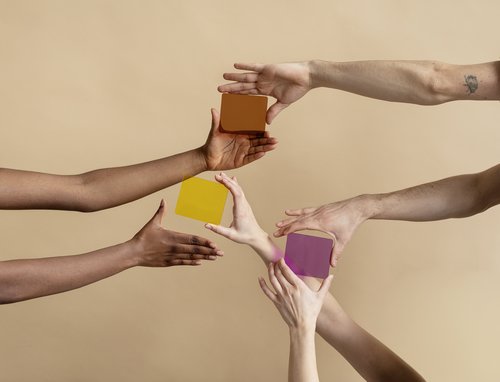 Verschiedene Hände greifen nach bunten, geometrischen Formen in Gelb, Lila und Orange vor einem neutralen Hintergrund.
