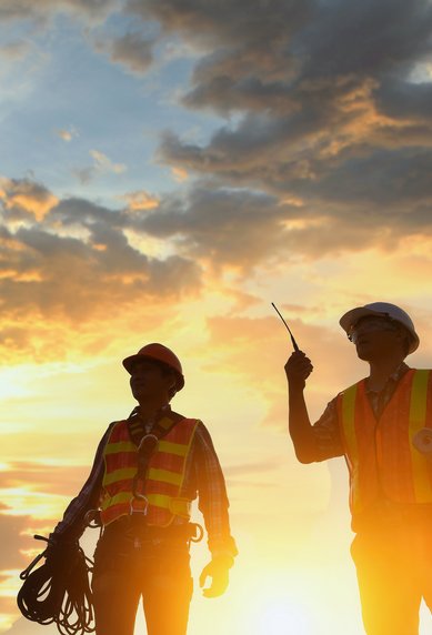 Two construction workers in safety vests stand at sunset, one holding a cable and the other pointing with a tool.