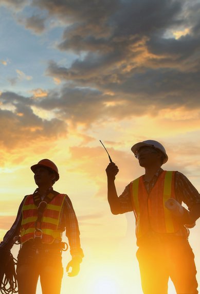 Two construction workers in safety vests stand at sunset, one holding a cable and the other pointing with a tool.