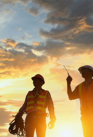 Zwei Bauarbeiter in Sicherheitswesten stehen bei Sonnenuntergang, einer hält ein Kabel und der andere zeigt mit einem Werkzeug.