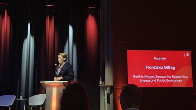 Franziska Giffey hält eine Keynote-Rede auf einer Veranstaltung. Im Hintergrund ist ein Bildschirm mit ihrem Namen und Titel als Bürgermeisterin von Berlin zu sehen. Die Bühne ist beleuchtet und es sind einige Zuhörer im Vordergrund.