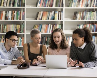 Four students are sitting at a table in a library, looking at a laptop. They are engaged in an animated discussion, with bookshelves visible in the background.