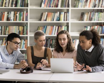 Vier Studierende sitzen an einem Tisch in einer Bibliothek und schauen auf einen Laptop. Sie diskutieren angeregt, während Bücherregale im Hintergrund sichtbar sind.