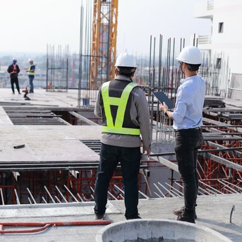 Two construction managers in hard hats are standing on a construction site, watching workers who are working on the structure of a building.