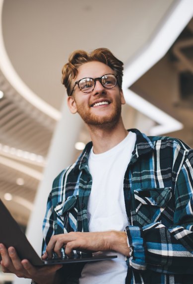 Young man with glasses and a checkered shirt, smiling as he holds a laptop in a modern interior.