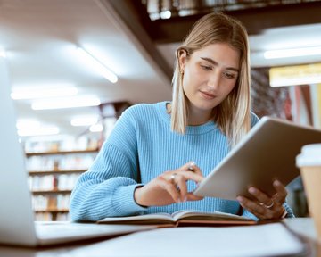 Eine junge Frau mit blonden Haaren sitzt in einer Bibliothek und nutzt ein Tablet. Vor ihr liegt ein aufgeschlagenes Buch, und neben ihr steht ein Kaffeebecher. Sie trägt einen blauen Pullover und konzentriert sich auf den Bildschirm.
