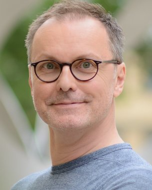 Ein Mann mit Brille und kurzem, grauem Haar lächelt in die Kamera, trägt ein graues T-Shirt und steht vor einem unscharfen Hintergrund.