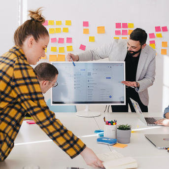 Ein Team von vier Personen arbeitet in einem modernen Konferenzraum. Eine Person präsentiert an einem Bildschirm, während die anderen aufmerksam zuhören und Notizen machen. Bunte Haftnotizen sind an der Wand angebracht.