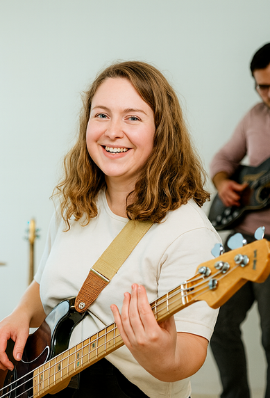 Eine Musikerin spielt Bass, während eine Sängerin am Mikrofon steht. Im Hintergrund sind ein Schlagzeuger und ein Gitarrist zu sehen.