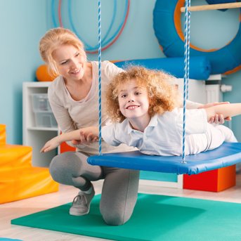 Eine Frau unterstützt ein Kind, das auf einer Schaukel liegt und lächelt. Der Raum ist mit Spielgeräten und Matten ausgestattet.