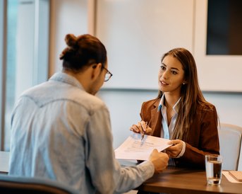 Eine Frau in einem Anzug überreicht einem Mann Dokumente während eines Gesprächs in einem modernen Büro.