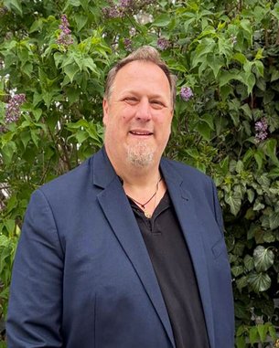 A man with a beard is wearing a dark blue blazer and a black T-shirt, standing in front of a green backdrop of plants with purple flowers.