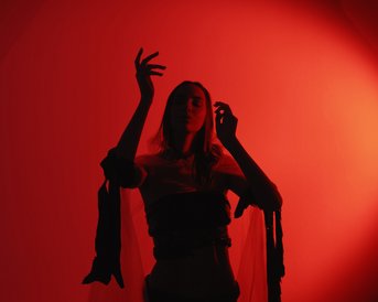 A person stands in red light, with raised hands and a mysterious expression, surrounded by a sheer fabric.
